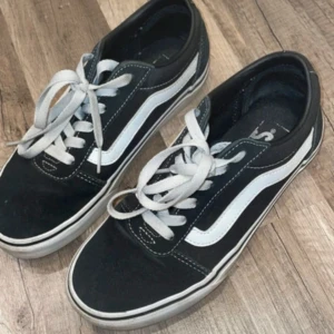 Svarta Vans Old Skool sneakers - Klassiska Vans Old Skool sneakers i svart med vit sidorand och vita snören. Skorna har låg profil, rund tå och platt sula. Ovandelen är i canvas och mocka med kontrasterande sömmar. Perfekta för en streetwear-look.