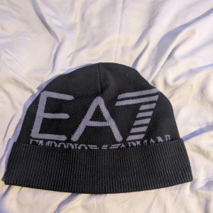 Svart EA7 Emporio Armani mössa - Svart mössa från EA7 Emporio Armani med stor grå logga framtill. Klassisk ribbad kant och mjukt stickat material som håller dig varm. Snygg och enkel design som passar perfekt till vintern. Skriv för frågor och bilder (: