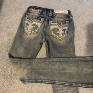 Rock Revival jeans med detaljer - Snygga blå Rock Revival jeans med slitningar och broderade bakfickor med silverfärgade detaljer och nitar. Jeansen har bootcut-modell och coola kontrastsömmar. Märkeslogga på bakfickan och midjebandet. Perfekta för dig som gillar statement-jeans.