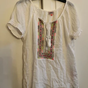 Vit blus med broderi från ONLY - Säljer en vit blus från ONLY i storlek 36. Blusen har korta ärmar, färgglatt broderi framtill i rosa, grön och svart samt snörning med tofsar vid halsen. Materialet är lätt och luftigt, perfekt för varma dagar. Snygg och bohemisk vibe!