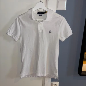 Vit pikétröja från Polo Ralph Lauren - Klassisk vit pikétröja från Polo Ralph Lauren med korta ärmar och krage. Tröjan har två knappar vid halsen och loggan i marinblått på bröstet.