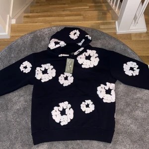 Blommig hoodie från Denim Tears - Säljer en mörkblå hoodie från Denim Tears med stora vita blommönster över hela tröjan. Hoodien har huva och magficka, samt en cool 'Made in USA'-patch. Perfekt statement-plagg för dig som gillar streetwear och unika prints.
