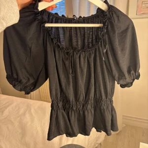 Svart blus med puffärmar och volang - Säljer en svart blus med puffiga korta ärmar och volang nedtill. Blusen har en rundad hals med snörning och smock i midjan för en snygg siluett. Materialet är mjukt och lätt samt kommer från Gina Tricot! Använd 1 gång🖤