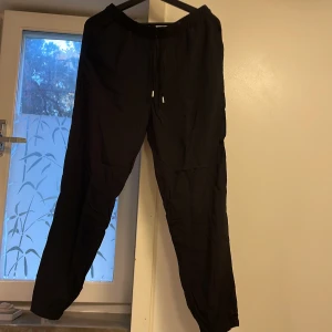 Nya svarta byxor från New Yorker - Svarta byxor från New Yorker med elastisk midja och snörning. Byxorna har resår vid bensluten för en avslappnad look och är tillverkade i ett lätt och mjukt material. Perfekta för chill dagar eller när du vill ha det bekvämt.