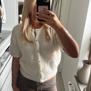 Vit kortärmad kofta från Gina Tricot - Supermysig vit kofta från Gina Tricot med korta ärmar och rund hals. Den är stickad i fluffigt material och har silvriga knappar framtill. Perfekt att slänga på sig för en chill look och funkar lika bra till jeans som till kjol.