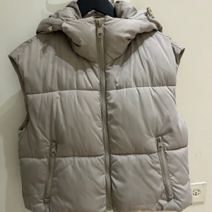 Ny Beige dunväst med huva från Zara - Snygg beige väst  från Zara i storlek XS. Västen har en quiltad design, praktisk huva och två fickor med dragkedja framtill. Perfekt för lager-på-lager och har en clean look som funkar till många stilar. Stängs med dragkedja framtill.