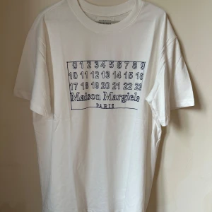 Maison Margiela vit t-shirt med print - Vit t-shirt från Maison Margiela med ikoniskt sifferprint och logga på bröstet. Klassisk rund hals och korta ärmar. Tillverkad i mjuk bomull, perfekt för dig som gillar minimalistisk design med en twist. Made in Italy.