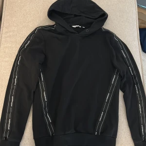 Svart hoodie från Antony Morato S - Svart hoodie från Antony Morato i storlek S med huva och långa ärmar. Snygga vita logoremsor längs ärmarna ger en sportig vibe. Tröjan har ribbade muddar och är tillverkad i mjuk bomullsmix som är skön att bära. Perfekt för chill eller streetwear.