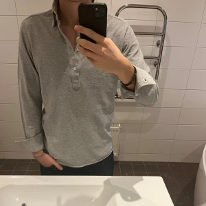 Långärmad pikétröja från John Henric - Säljer en stilren grå långärmad piké från John Henric. Tröjan har lång ärm, klassisk krage och knappar framtill. Som ny och riktigt snygg. 