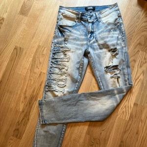 AMIRI jeans - Säljer ett par ljusblå slim fit jeans från AMIRI med stora slitningar och broderad AMIRI-logga längs benet. Jeansen har klassisk femficksdesign, dragkedjegylf och coolt distressed utseende. Perfekta för dig som gillar streetstyle och detaljer. Helt nya! Riktiga!