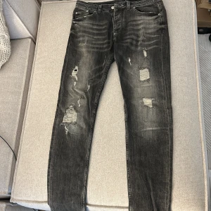 Svarta distressed jeans från Dsquared2 - Säljer ett par svarta jeans från Dsquared2 i storlek 50. Jeansen har slitna detaljer och trasiga partier framtill för en riktigt edgy look. De är raka i modellen och har klassisk femficksdesign. Perfekta för dig som gillar streetstyle och vill sticka ut.