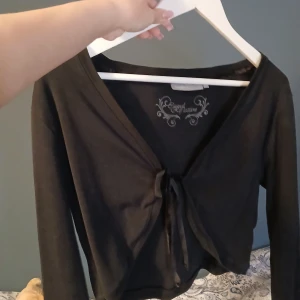 Svart crop kofta 2000s stil - Säljer en svart croppad bolerotopp från Easy Collection. Toppen har långa ärmar, v-ringning och knyt framtill. Perfekt att slänga över ett linne eller klänning för en snygg lager-på-lager-look. Enkel och stilren design som funkar till många outfits.