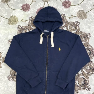 Marinblå hoodie från Polo Ralph Lauren - Snygg och klassisk mörkblå zip up hoodie från Polo Ralph Lauren. Tröjan har huva med vita snören, fickor framtill och den ikoniska gula loggan broderad på bröstet.  Längd från axeln och ner: 59 cm Bredd under armhålorna: 49 cm  📦 Skickas snabbt & välpackat.