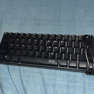 MageGee TS91 Membrane Keyboard - Kompakt MageGee TS91 membrantangentbord med 61 tangenter. Perfekt för gaming eller arbete där platsen är begränsad. Synligt damm men i gott skick, inga uppenbara skador. Ansluts via kabel, DC 5V. Snygg design med unikt logotryck på mellanslagstangenten.