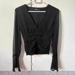 Svart v-ringad blus från BikBok  - Säljer en svart blus från BikBok i storlek XS. Blusen har v-ringning, långa transparenta ärmar och rynkade detaljer framtill. Modellen är croppad och har smock baktill för en snygg passform. Perfekt för dig som gillar stilrena och trendiga plagg. Finns en fläck på armen en mindre fläck som syns på sista bilden!🤗❤️