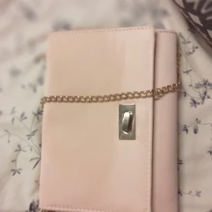 Ljusrosa kuvertväska med guldkedja - Snygg ljusrosa kuvertväska med stilren guldfärgad kedja och metallspänne framtill. Väskan har en slät yta och är perfekt att bära över axeln. Insidan är fodrad i matchande rosa tyg och har ett litet innerfack för småsaker.