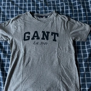 Grå GANT t-shirt med tryck - Snygg grå t-shirt från GANT med stort svart tryck på bröstet där det står 'GANT Est. 1949'. Klassisk rund halsringning med randig detalj på insidan av kragen. Perfekt basic-plagg med stilren look.