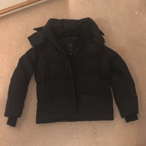 Canada Goose Wyndham - Säljer en svart pufferjacka från Canada Goose i storlek S. Jackan har hög krage, dragkedja och tryckknappar framtill samt ribbade muddar vid ärmsluten. Klassisk patch på ärmen och riktigt varm för kalla dagar. Kommer med päls. NFC Scan✅