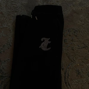 Svarta Juicy Couture mjukisbyxor - Svarta mjukisbyxor från Juicy Couture i mjuk sammet med glittrande strasstext på baksidan och ett stort J på framsidan. Byxorna har dragsko i midjan och klassisk bootcut-modell. Perfekta för en chill och trendig look.