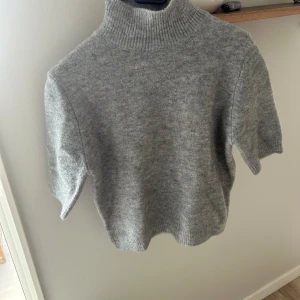 Grå stickad polotröja med kort ärm - Supermjuk och stilren grå stickad polotröja med korta ärmar och ribbad hög krage. Perfekt till jeans eller kjol för en clean look. Tröjan har en något croppad passform och är gjord i ett mysigt material som känns lyxigt mot huden.