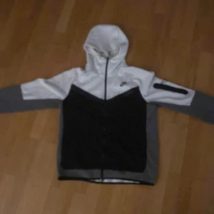 Nike vindjacka med huva, svart/vit/grå - Nike vindjacka i svart, vitt och grått med hel dragkedja och huva. Jackan har lång ärm, en ficka med dragkedja på ena ärmen och klassisk Nike-logga på bröstet. Perfekt för dig som gillar sportig och clean stil.