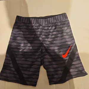 Nike Dri-Fit mönstrade träningsshorts - Nike Dri-Fit träningsshorts i slim fit med vågigt mönster i mörkgrått och svart. Snygg orange/röd swoosh på benet och elastisk midja för skön passform. Tillverkade i 100% polyester, perfekta för sport och aktivitet.