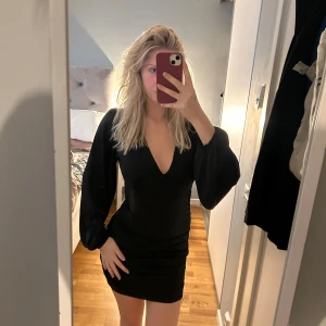 Svart kort klänning med v-ringning - Säljer en svart kort klänning med djup v-ringning och långa puffärmar. Klänningen har en tight passform som framhäver figuren och är perfekt för dig som gillar en stilren och trendig look. Materialet känns mjukt och följsamt mot kroppen.