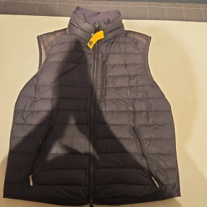 Svart dunväst från Parajumpers S - Snygg svart dunväst från Parajumpers i storlek S. Västen har quiltad design, hög krage och dragkedja framtill samt praktiska fickor med dragkedja. Detaljer i gult och klassisk Parajumpers-logga i nacken. Perfekt för lager-på-lager och streetstyle. Den har lite lagningar till exempel på en av dragkedjorna.men annars är den i bra skick.