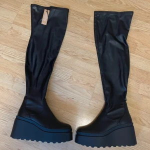 Svarta overknee boots med platå - Säljer ett par svarta overknee boots med hög platåsula och rund tå. Skorna är i syntetmaterial med stretchig skaft som går över knäet. Perfekt skick, aldrig använt tyvärr! Köpta för drygt 900kr. 