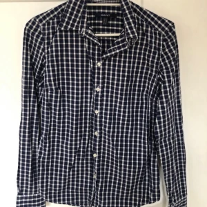 Blårutig skjorta från GANT - Snygg blårutig skjorta från GANT med klassisk krage och vita knappar framtill. Skjortan har lång ärm och är tillverkad i mjuk bomull som känns skön mot huden. Perfekt för dig som gillar stilrena och tidlösa mönster.