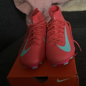 Nike Mercurial rosa fotbollsskor - Säljer ett par Nike Mercurial fotbollsskor i en riktigt snygg rosa färg med turkosa detaljer och logga. Skorna har snörning, högre strumpliknande skaft och plös, samt dobbar i turkos och rosa för bästa grepp på planen. Perfekta för dig som vill sticka ut på fotbollsplanen helt nya.