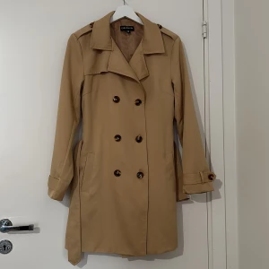 Beige trenchcoat från EllyBazar XS - Stilren beige trenchcoat från EllyBazar i storlek XS. Jackan har dubbelknäppning, klassisk krage, bälte i midjan och snygga knappar på ärmsluten. Perfekt för dig som vill ha en tidlös look med lite edge.
