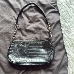 Svart axelväska med kedjedetaljer - Snygg svart axelväska med croco-mönstrad yta och silverfärgade kedjedetaljer på axelremmen. Väskan har dragkedja upptill och ett rymligt fack med innerficka. Perfekt storlek för det viktigaste och ger en cool vibe till din outfit.