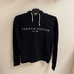 Svart hoodie Tommy Hilfiger XS - Svart hoodie från Tommy Hilfiger i storlek XS, tillverkad av ekologisk bomull. Tröjan har vit snörning i huvan, broderad logga och ficka framtill. Snygg och enkel design med lång ärm och klassisk passform.