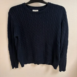 Mörkblå stickad tröja Holly & White - Snygg mörkblå stickad tröja från Holly & White med klassiskt kabelstickat mönster. Rund hals och långa ärmar ger en chill och tidlös vibe. Perfekt för dig som gillar enkel men stilren look.