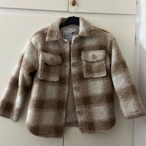 Rutig teddyjacka från Skill, beige/vit - Supermjuk teddyjacka från Skill i rutigt beige och vitt. Jackan har två stora bröstfickor med knappar, klassisk krage och stängs med knappar framtill. Perfekt för kalla dagar när du vill vara både varm och snygg.