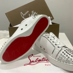 Christian Louboutin vita sneakers med nitar - Snygga vita sneakers från Christian Louboutin med ikonisk röd sula och coola silverfärgade nitar på tå och häl. Skorna har snörning och är tillverkade i skinn med detaljer i silver. Perfekta för dig som vill sticka ut med en lyxig och edgy stil.
