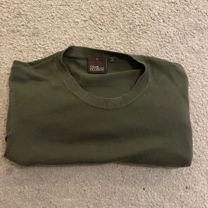 Mörkgrön stickad tröja Oscar Jacobson - Snygg mörkgrön stickad tröja från Oscar Jacobson, tillverkad i Italien. Tröjan har rund halsringning, långa ärmar och ribbade muddar vid hals, ärmslut och nederkant. Perfekt för dig som gillar stilrena och klassiska plagg. Obs de har ett litet hål i ryggen.