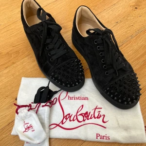 Christian Louboutin svarta - Svarta sneakers från Christian Louboutin i mocka med ikoniska röda sulor och svarta nitar på tån. Skorna har snörning och rund tå, samt exklusiva detaljer som ger en edgy och lyxig vibe. Perfekta för dig som vill sticka ut med stil. Storlek 42. Kvitto finns! Priset kan diskuteras 👌