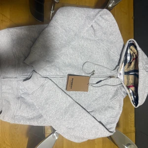 Grå hoodie från Burberry med dragkedja - Snygg grå hoodie från Burberry med klassisk rutig insida i huvan. Tröjan har dragkedja framtill, snörning vid huvan och ribbade muddar. Perfekt för dig som gillar stilrena detaljer och exklusiva märken.