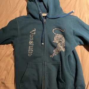 Mörkblå hoodie med paljett-tiger - Snygg mörkblå hoodie från Vonstudio med dragkedja och huva. Framsidan har ett coolt paljettmotiv av en tiger och texten 'Vonstudio' i glittriga paljetter. Tröjan har långa ärmar, fickor och snörning vid huvan. Perfekt för dig som gillar statement-plagg. 