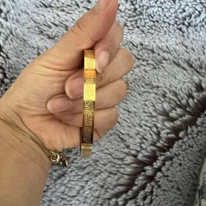 Guldigt armband med gravyr, ayatul kursi - Snyggt armband i guldfärgad metall med detaljerad gravyr i form av arabisk text runt hela bandet. Armbandet är stelt och har en minimalistisk, stilren design som passar perfekt för dig som gillar unika accessoarer med personlig touch.