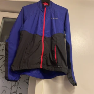 Lilla och svart vindjacka från SOC - Sportig vindjacka från SOC i lilla och svart med rosa dragkedjor. Jackan har långa ärmar och två fickor framtill. Tillverkad i lätt polyester med meshfoder för extra komfort. Perfekt för utomhusaktiviteter när det blåser.