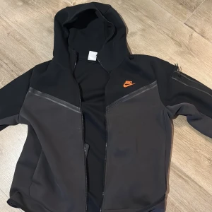 Svart hoodie från Nike med dragkedja - Svart hoodie från Nike med orange logga på bröstet. Tröjan har hel dragkedja, huva och praktiska fickor. Materialet är mjukt och sportigt, perfekt för chill eller träning. Snygga detaljer med svarta paneler och diskret mönster på ärmen.