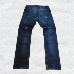 Vintage Jack & Jones distressed Archive bootcut wide loose washed blue Y2K jeans rare grisch niche - Säljer ett par coola Jack & Jones Originals Tim Slim Fit jeans i storlek W32 med en snygg mörkare tvätt och fabriksgjorda slitningar. Dessa jeans har en klassisk och bekväm slim fit-passform i god, robust denimkvalitet. Viktigt att veta är att det finns ett litet hål i sömmen på baksidan vid grenen. Detta kräver en enkel reparation för att byxorna ska bli perfekta igen, vilket reflekteras i priset. Ett utmärkt kap för den händige som vill ha ett par snygga märkesjeans! Skicka dm för mått :)