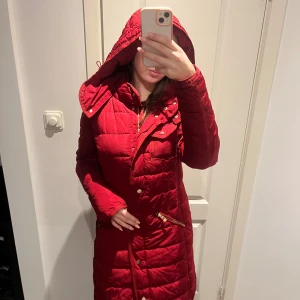 Röd lång pufferjacka från Desigual - Lång röd pufferjacka från Desigual med snygg quiltad yta, guldiga knappar och dragkedja. Jackan har huva och ett unikt blommigt innerfoder med färgglada detaljer. Perfekt för kalla dagar och sticker ut med sin coola design.