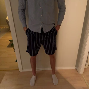 Randiga shorts från Kappahl, marinblå - Snygga marinblå shorts från Kappahl med vita vertikala ränder och elastisk midja med snörning. Shortsen har fickor på sidorna och en bakficka, perfekt för en chill sommardag. Lätta och sköna att bära.