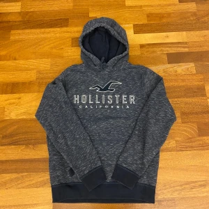 Mörkgrå hoodie från Hollister - Mysig mörkgrå hoodie från Hollister med vit logga och text framtill. Tröjan har huva med snörning och ribbade muddar vid ärmslut och nederkant. Perfekt för chill dagar och snygg till jeans eller mjukisbyxor. Vintage och väldigt efterfrågatt plagg