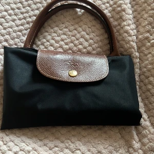 Svart Longchamp handväska - Klassisk svart handväska från Longchamp med bruna skinnhandtag och lock. Väskan har ett lock med guldfärgad knapp. Perfekt storlek för att bära med sig allt man behöver och enkel att vika ihop.