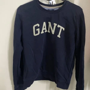 Mörkblå sweatshirt från GANT - Säljer en mörkblå sweatshirt från GANT med klassisk rund hals och stora vita bokstäver framtill. Tröjan är i mjuk bomull och har långa ärmar. Perfekt för en chill och stilren look.